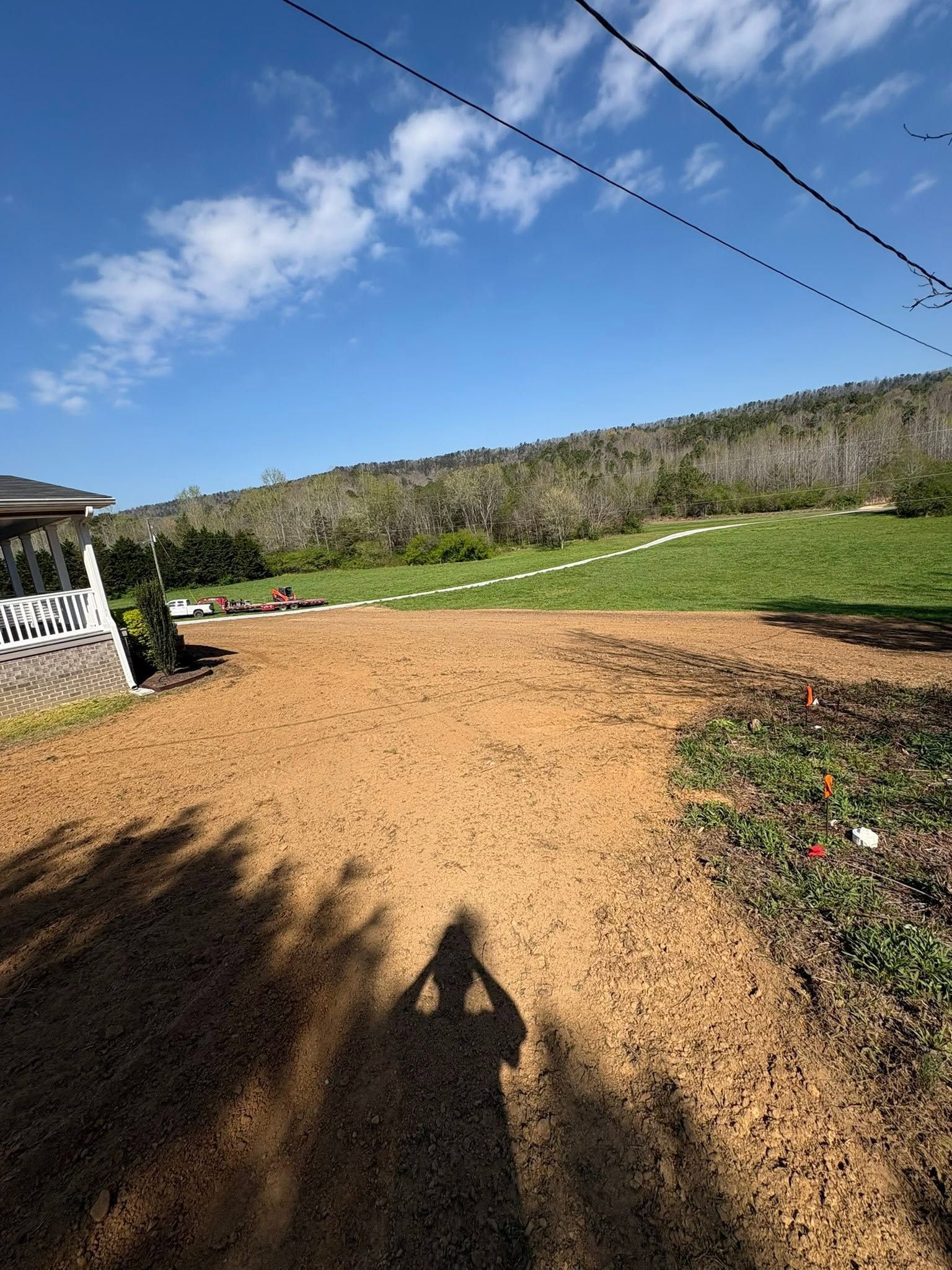 Dirt driveway beside a grassy field and tree line under a blue sky with power lines overhead.