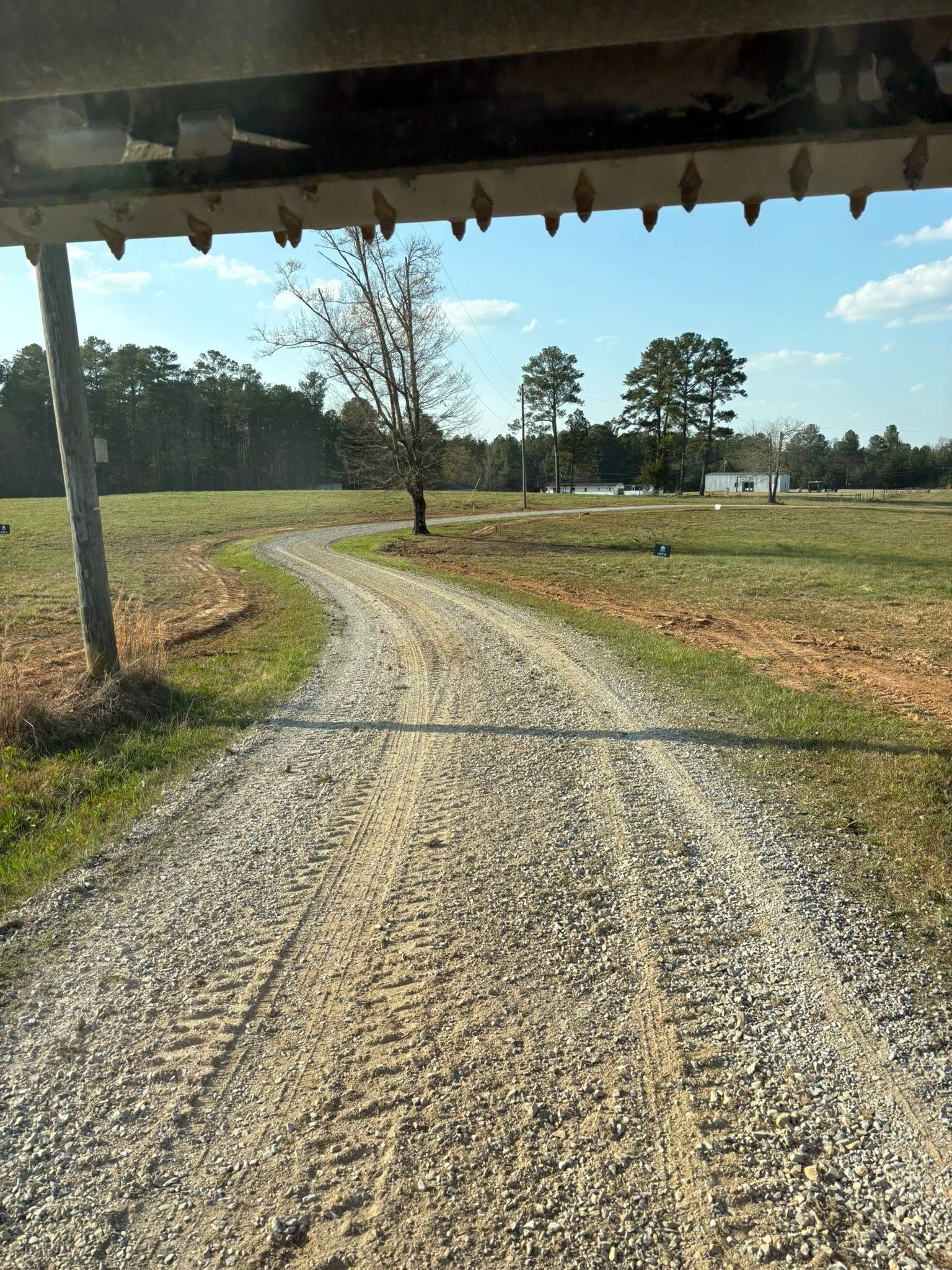 Gravel driveway curving through a grassy rural field under a clear blue sky, viewed from a shaded porch.