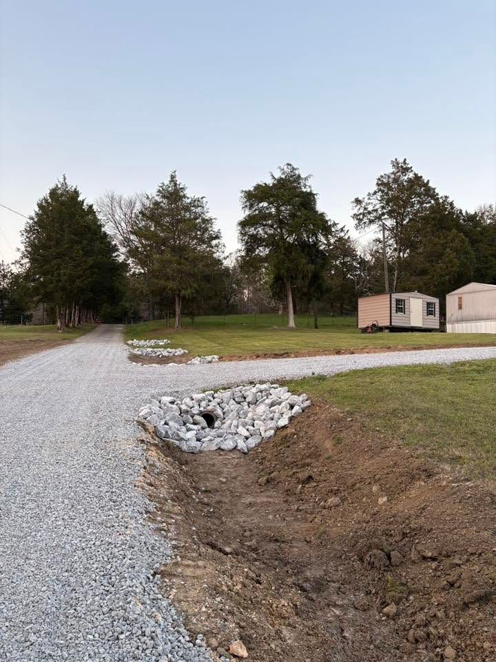Gravel driveway beside a grassy lot, with white rock-lined drainage trenches and small buildings in the background