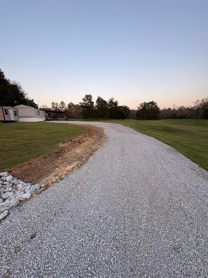 Gravel driveway curving through a grassy field at dusk, with trees and a small camper on the left.