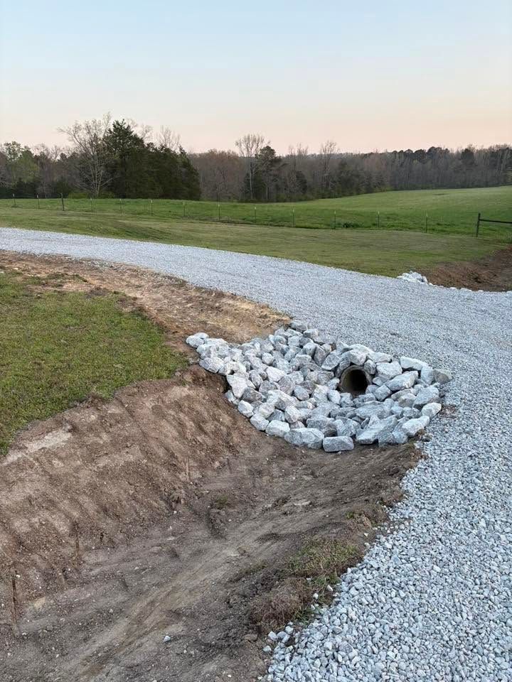 Gravel driveway with a culvert drainage pipe and white riprap beside a grassy field at dusk