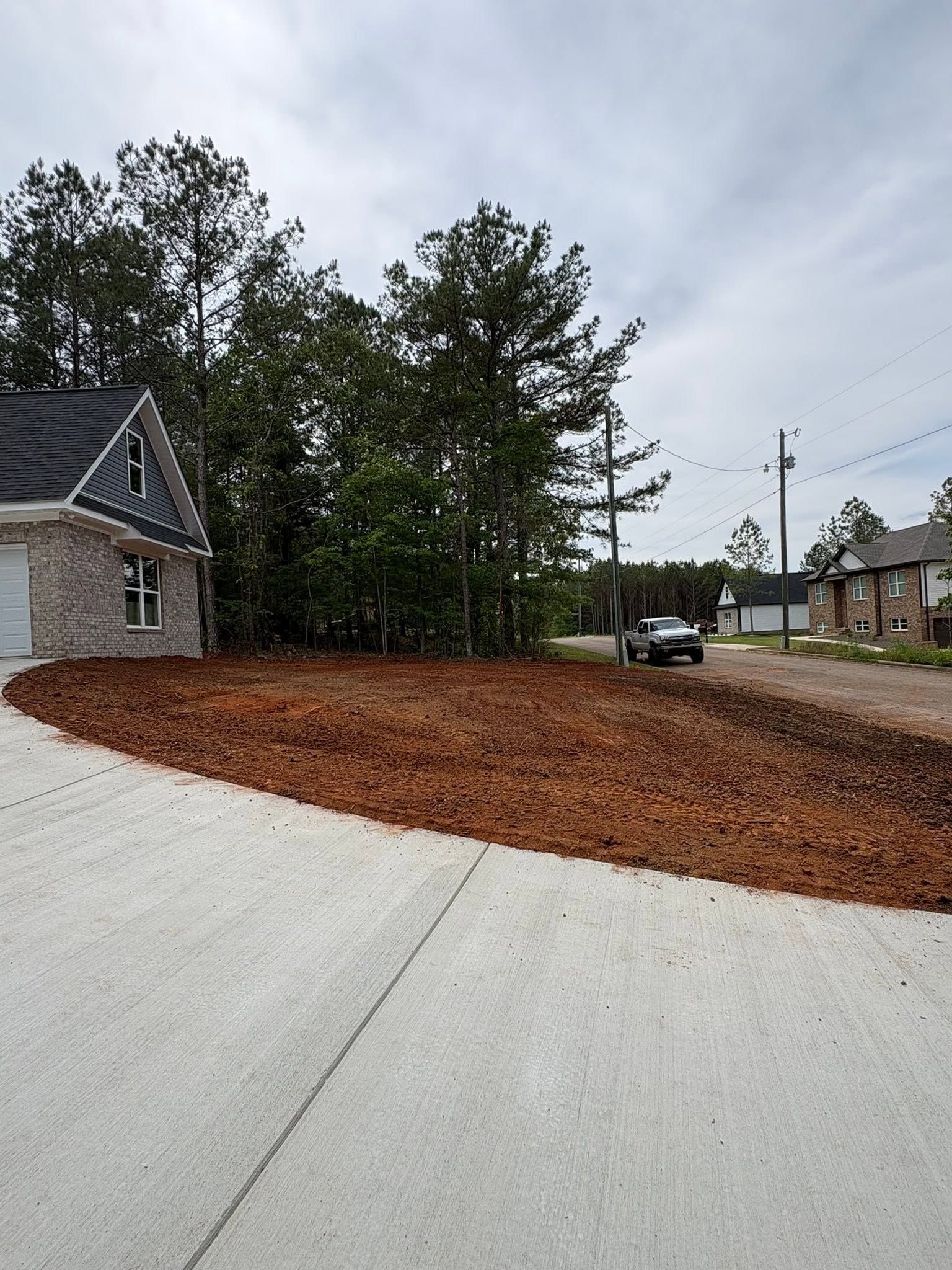 Curved concrete driveway beside a mulch-covered slope, trees, and houses under an overcast sky