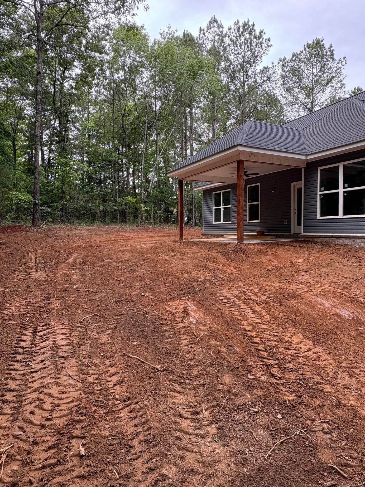 New house under construction with brown siding, covered porch, and muddy yard beside trees
