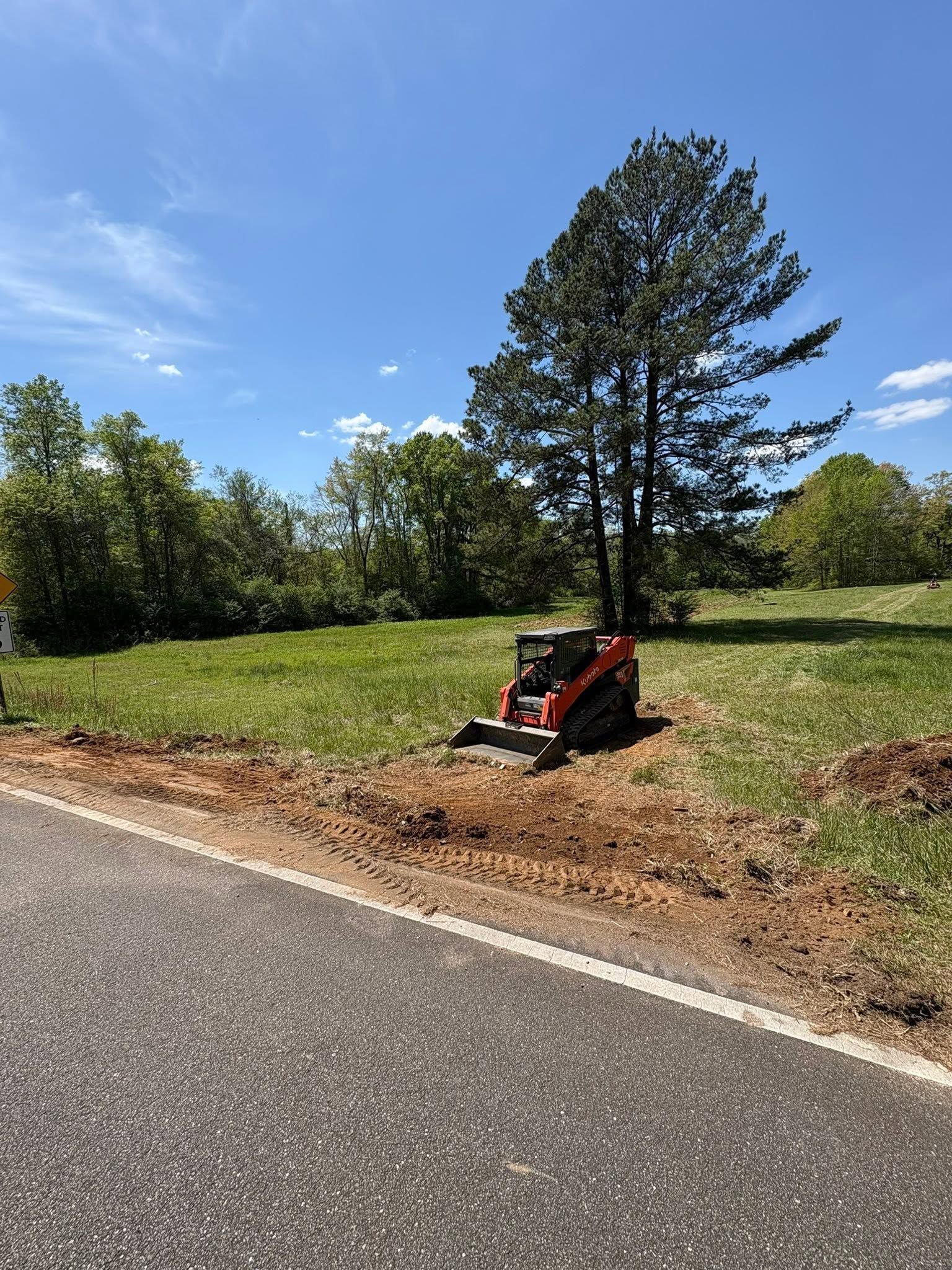 Red skid steer parked on a dirt shoulder beside a road under a blue sky.
