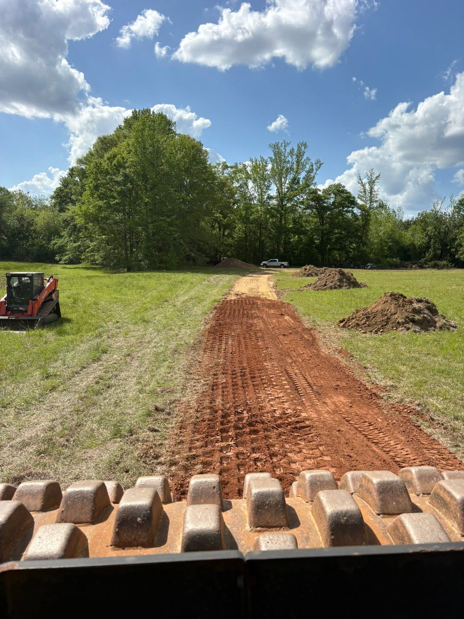 Brick pavers lined up in front of a dirt path leading across a grassy field under a blue, cloudy sky.