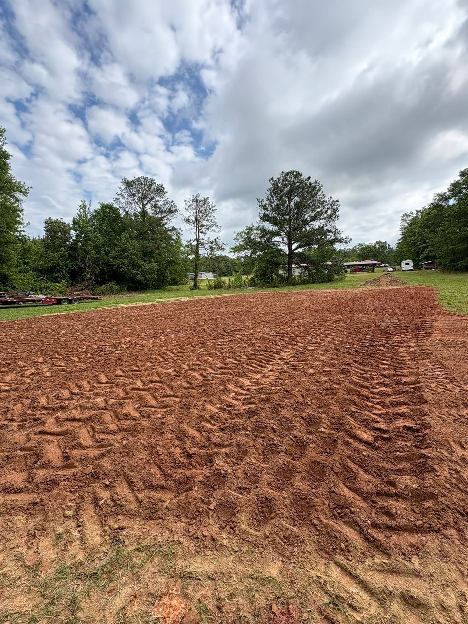 Freshly tilled red-brown field under a cloudy sky, with trees and a distant house on the horizon