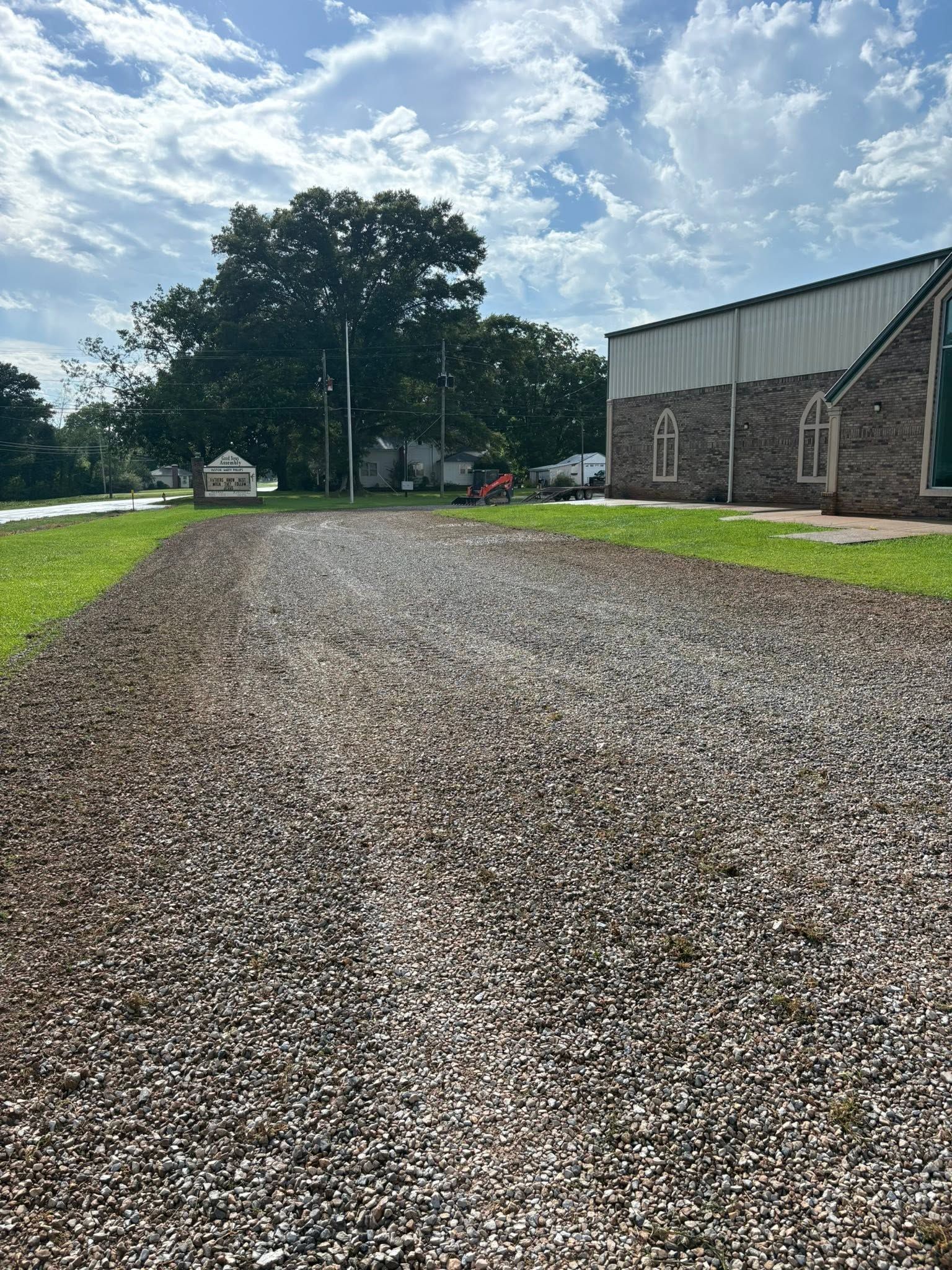 Gravel road beside a stone building under a partly cloudy sky, with trees in the distance