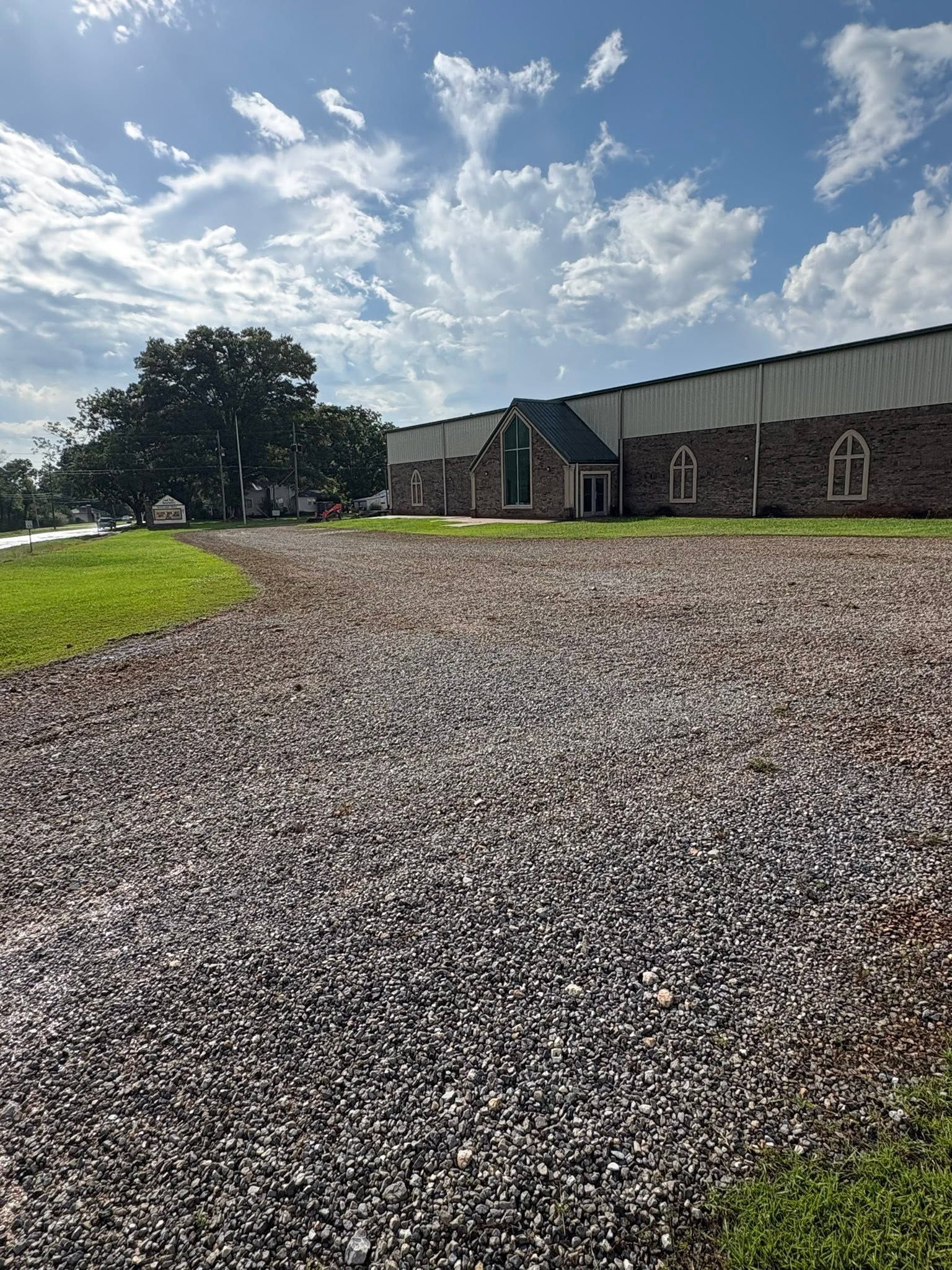 Gravel courtyard beside a brick building under a bright cloudy sky, with a large tree and green lawn.