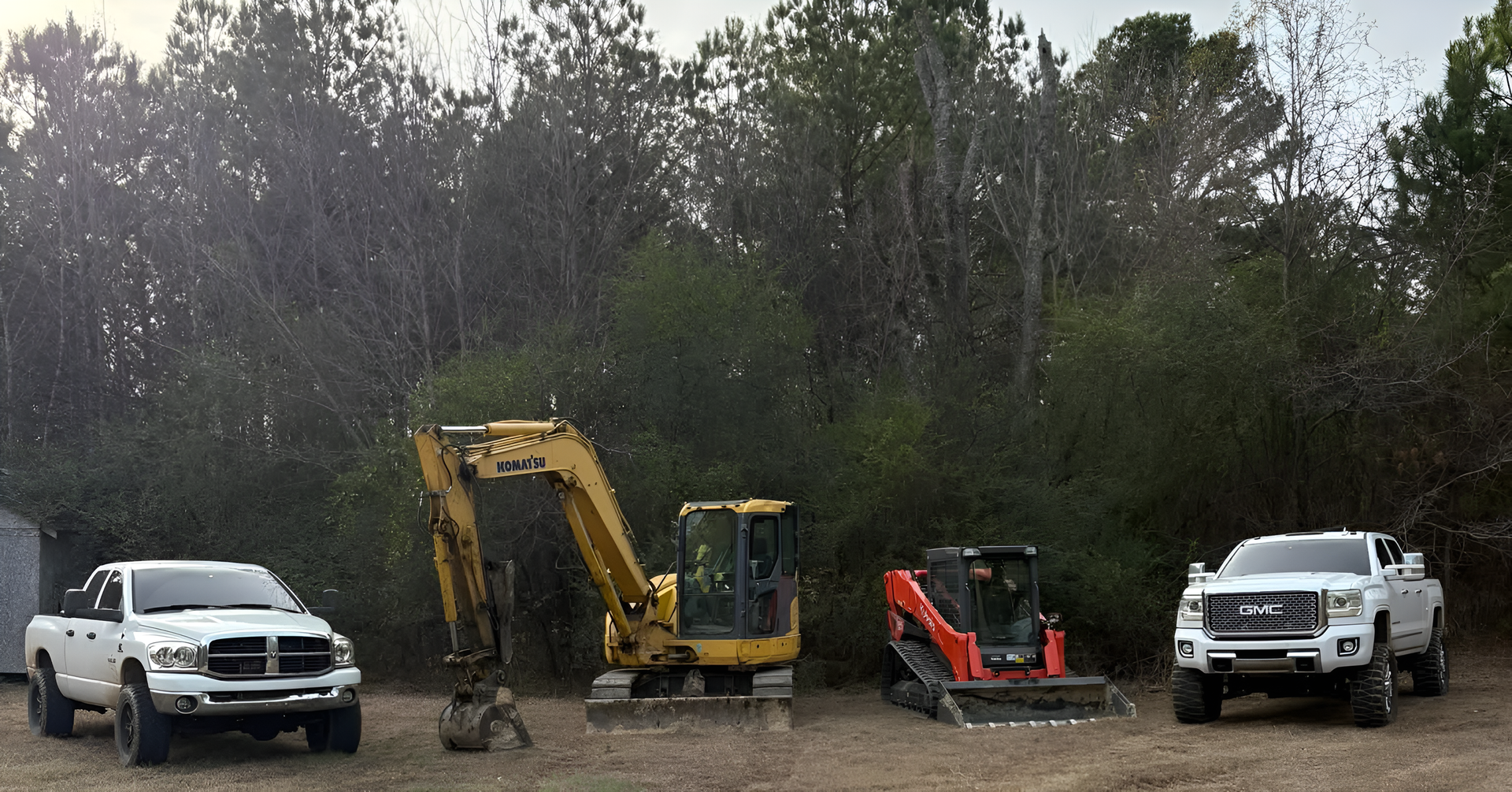 White pickup, yellow excavator, red skid steer, and white truck parked in a muddy lot during rain.
