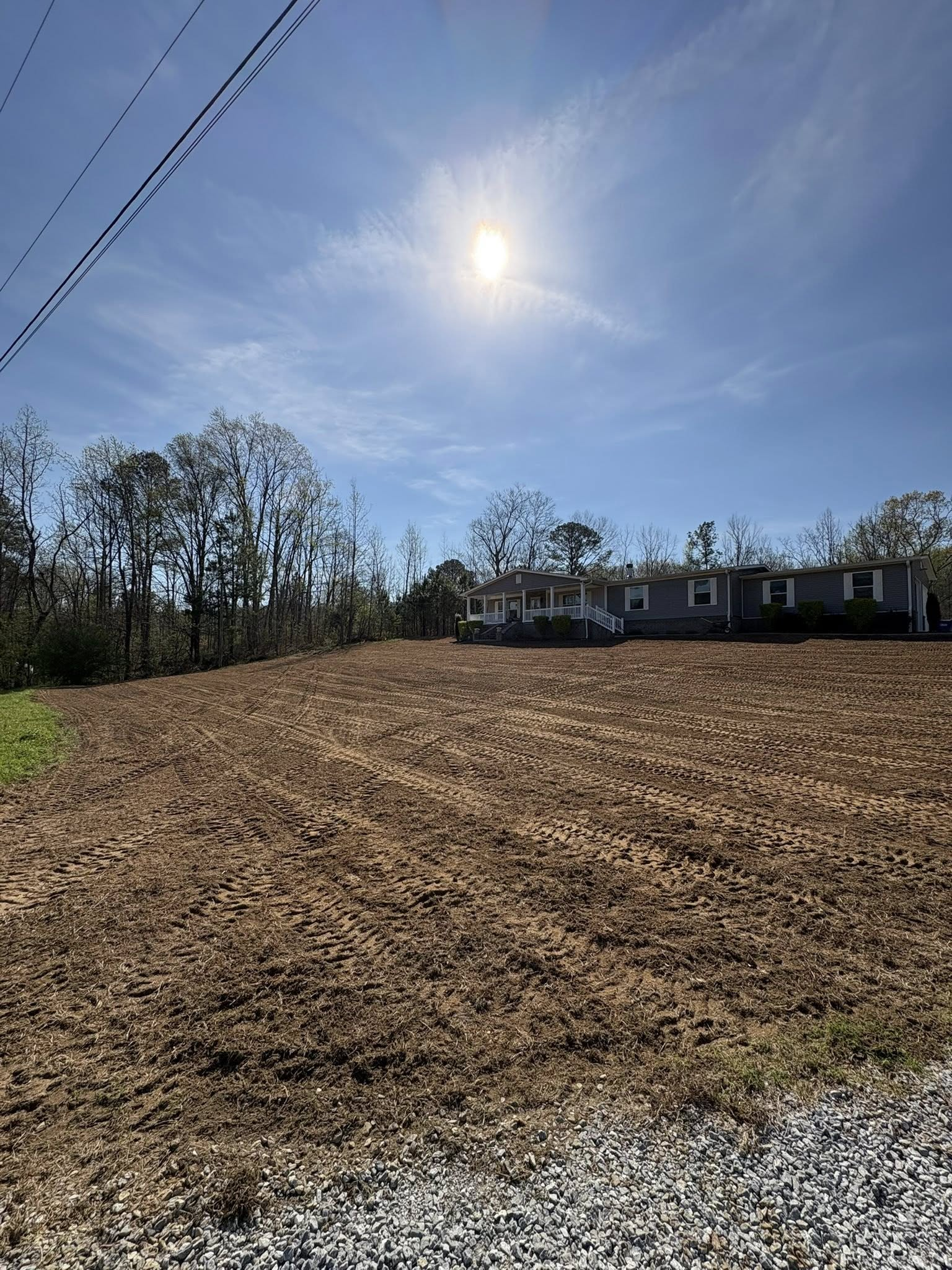 Plowed field under a bright sun, with trees and a house line in the distance.