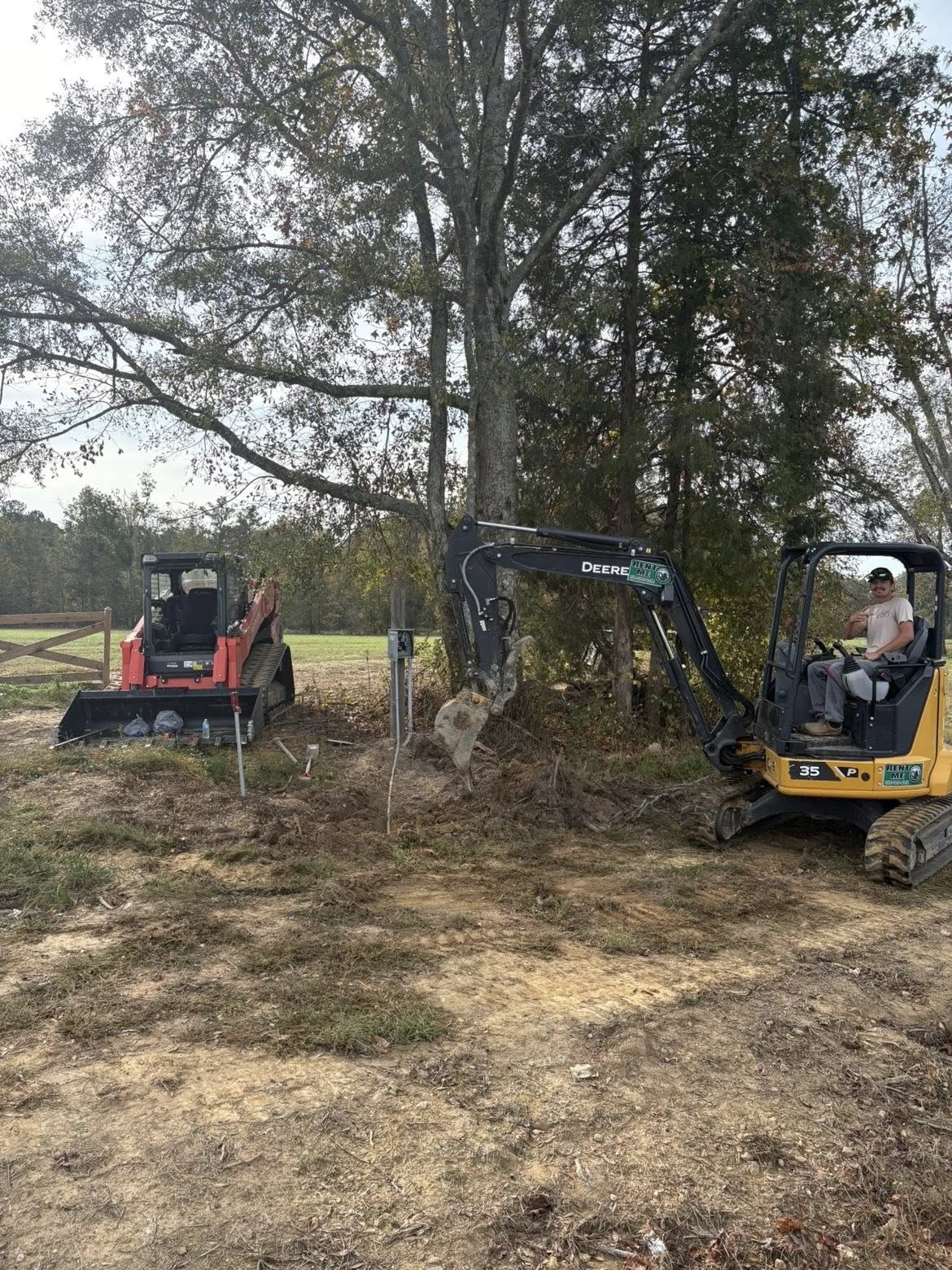 Two excavators working beside a tree in a grassy field, one red and one yellow.