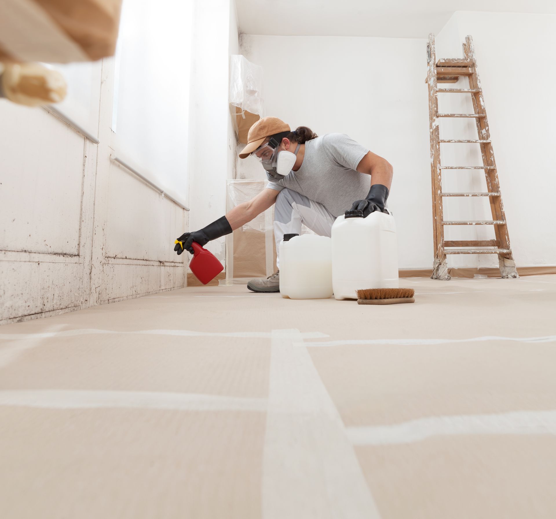 Person in a room with painting supplies, kneeling while spraying. Ladder and containers are nearby.