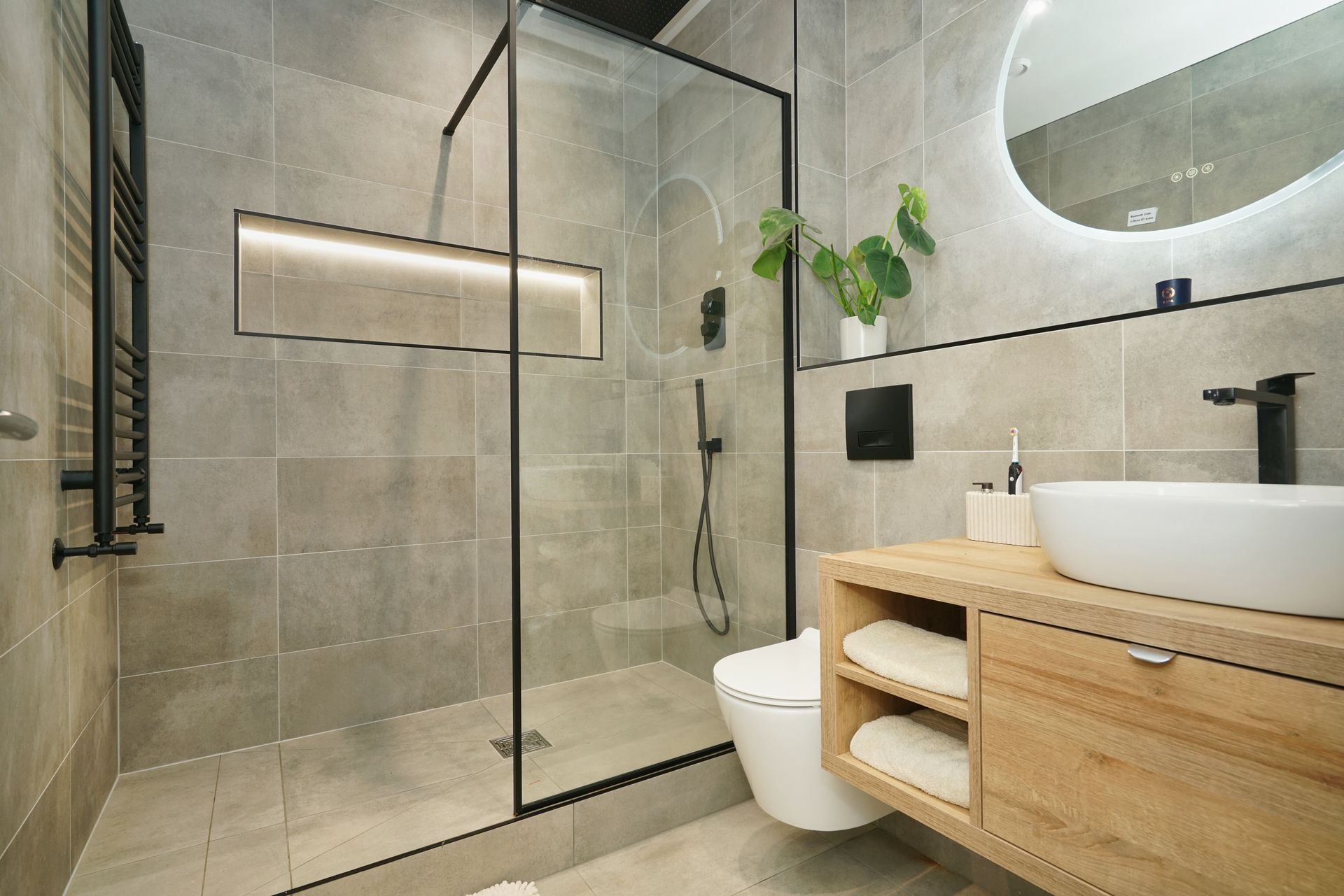 Modern bathroom with gray tiled walls, glass shower, wooden vanity, and round mirror.