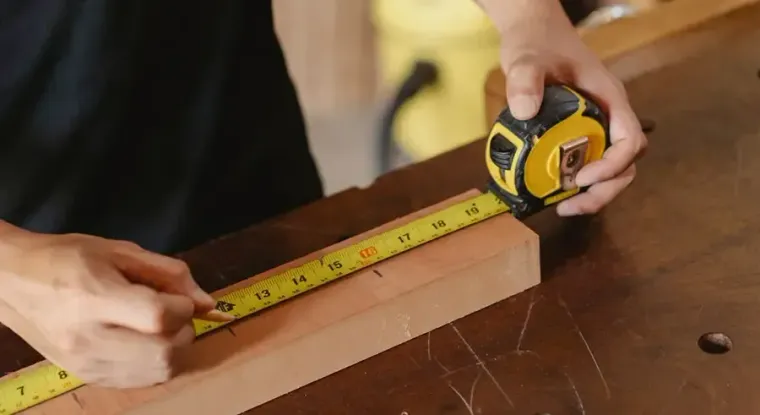 Hands measuring a wooden board with a yellow tape measure on a workbench