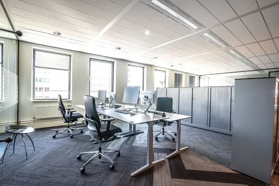 Empty modern office with desks, rolling chairs, partition screens, and large windows admitting daylight
