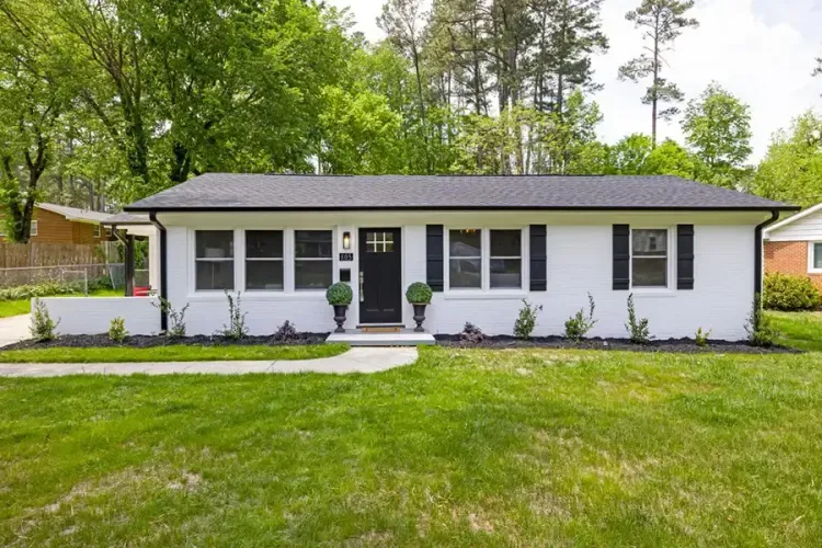 Single-story white house with black shutters, front porch, and a green lawn surrounded by trees