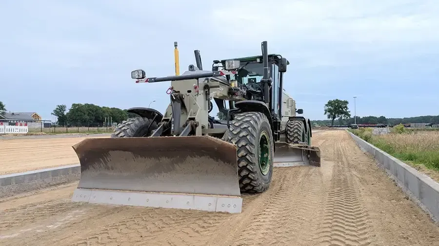 Gray bulldozer grading a sandy road beside concrete curbs under a cloudy sky
