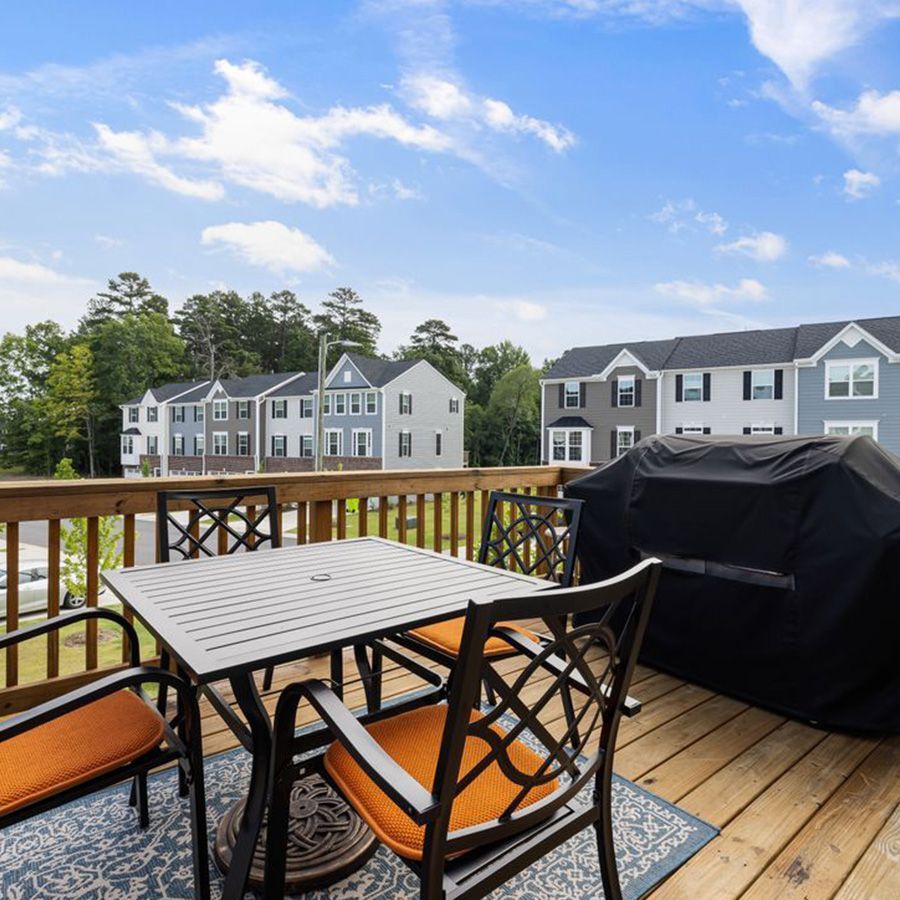 Wooden deck with table, chairs, and grill overlooking apartment buildings under a blue sky