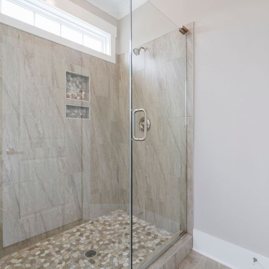 Glass walk-in shower with beige tile walls, pebble floor, and a chrome handle in a bright bathroom