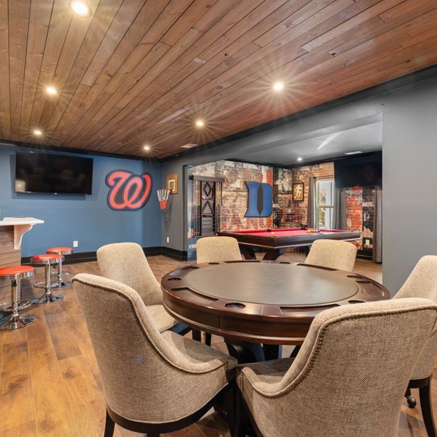 Cozy basement lounge with round table, beige chairs, bar stools, TV, and blue accent wall with neon signs