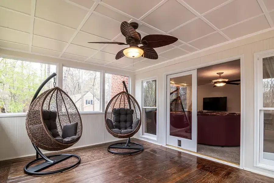 Covered porch with two hanging egg chairs, ceiling fan, and glass doors overlooking a wooded view