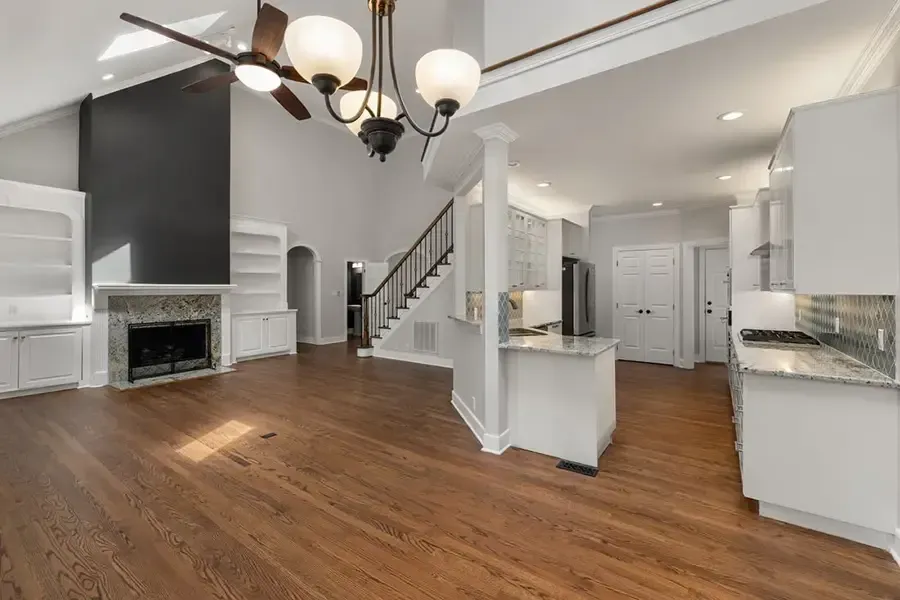 Bright open-plan living room with hardwood floors, white cabinets, fireplace, staircase, and black chandelier