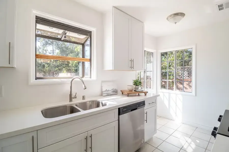 Bright white kitchen with sink, dishwasher, cabinets, and sunlit windows.
