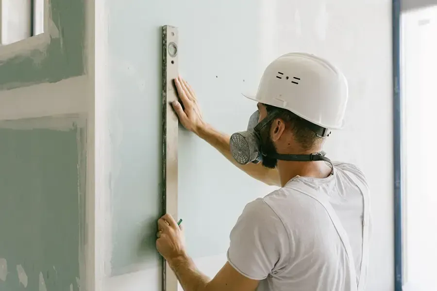 Worker in a white hard hat measuring a wall with a level during drywall installation.