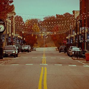 Empty autumn street with yellow center lines, parked cars, and storefronts under warm lights
