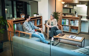 Two people sit in armchairs in a modern office, having a conversation across a coffee table with documents. Two people sit in armchairs in a modern office, having a conversation across a coffee table with documents.