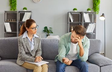 A professional sits with a notepad, observing a person with their head in their hand, looking distressed on a gray sofa. A professional sits with a notepad, observing a person with their head in their hand, looking distressed on a gray sofa.