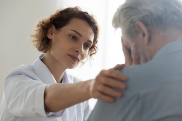 A healthcare provider in a white coat places a comforting hand on the shoulder of an upset person covering their face. A healthcare provider in a white coat places a comforting hand on the shoulder of an upset person covering their face.