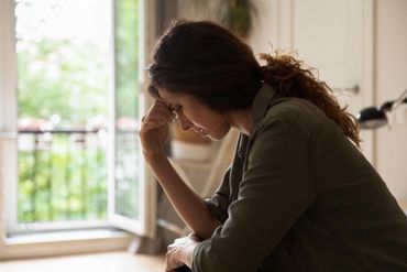 A person sits in profile, resting their forehead on their hand, looking down with a somber expression in a bright room. A person sits in profile, resting their forehead on their hand, looking down with a somber expression in a bright room.
