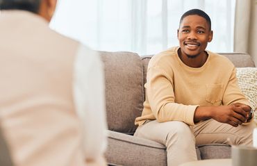 A person in a beige sweater sits on a grey sofa, smiling while speaking with someone off-camera in a bright room. A person in a beige sweater sits on a grey sofa, smiling while speaking with someone off-camera in a bright room.