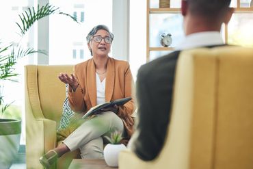 A professional in a tan blazer sits in a yellow chair, gesturing while speaking to someone seated across from them. A professional in a tan blazer sits in a yellow chair, gesturing while speaking to someone seated across from them.