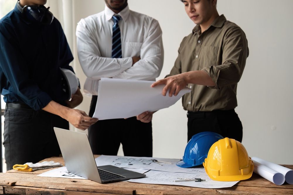 Three people review construction plans at a desk with a laptop, blueprints, and yellow hard hats.