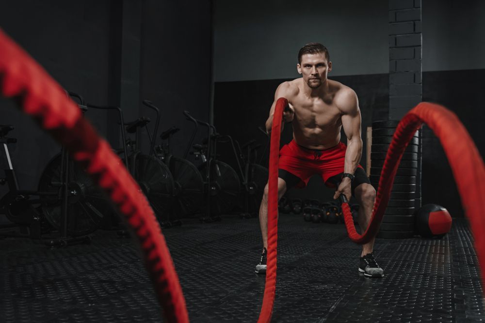 Man exercising with battle ropes in a dark gym, wearing red shorts and gloves