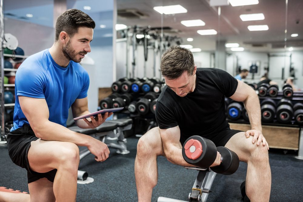 Two men exercising with dumbbells in a gym, one seated and one crouched, with weight racks behind them.
