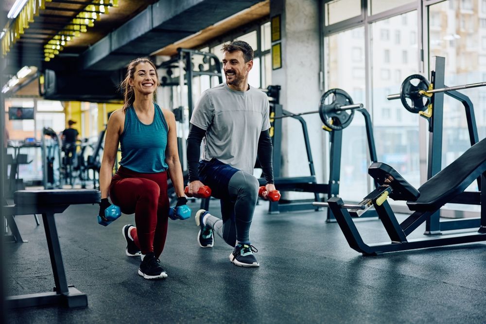 Two people lunging with dumbbells in a bright gym.