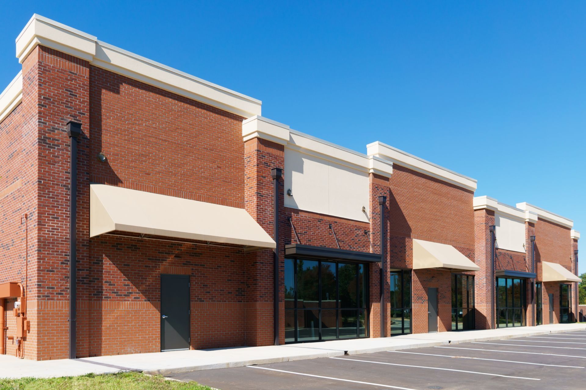 Row of empty red-brick storefronts with awnings and large windows on a sunny day