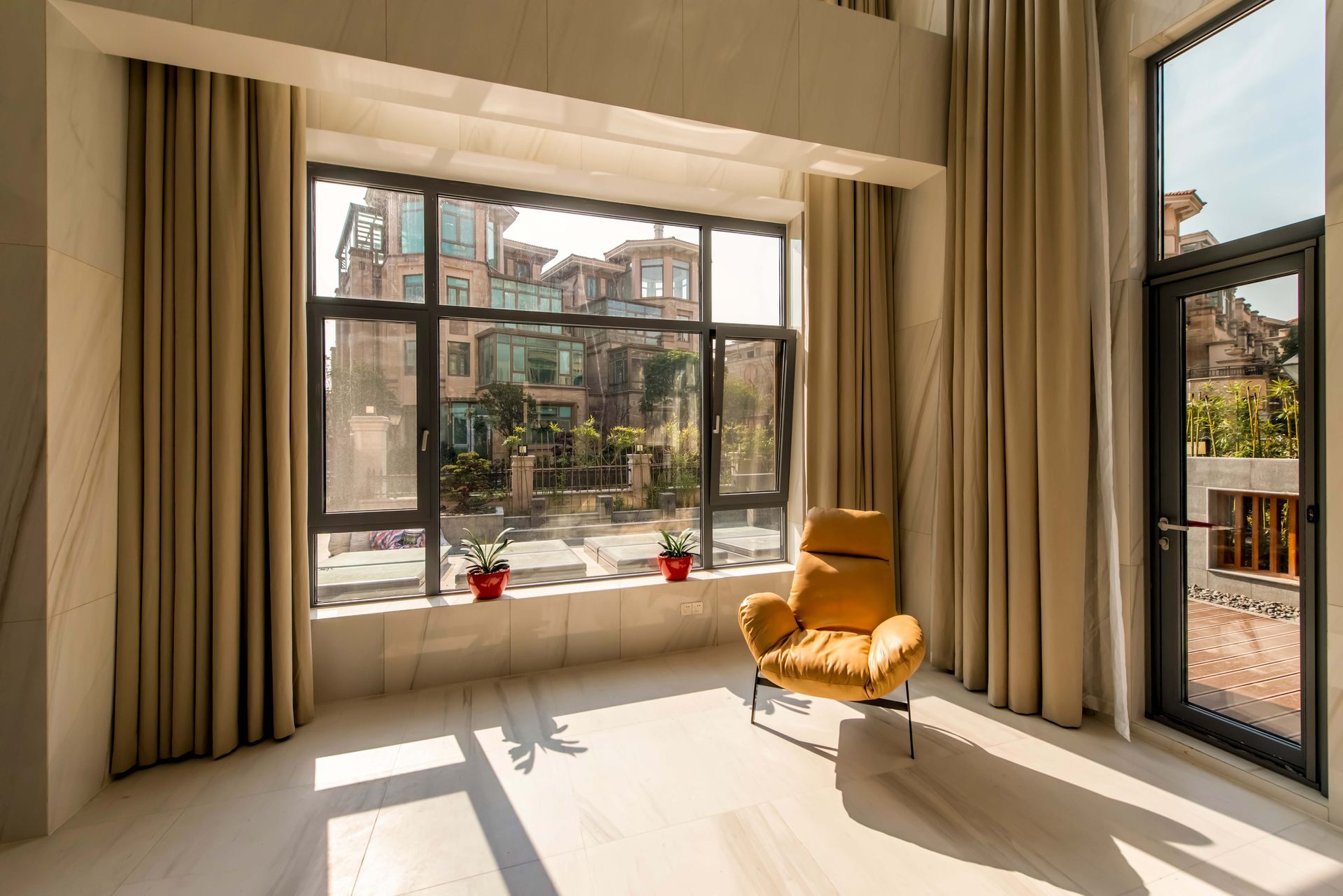 Bright sunlit room with beige curtains, large windows, and a mustard armchair by a glass door.