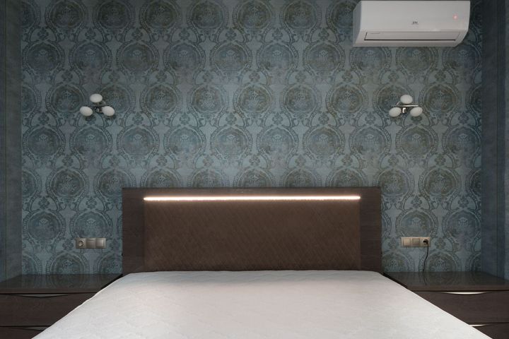 Modern bedroom with patterned teal wallpaper, white bed, dark headboard, wall lamps, and air conditioner