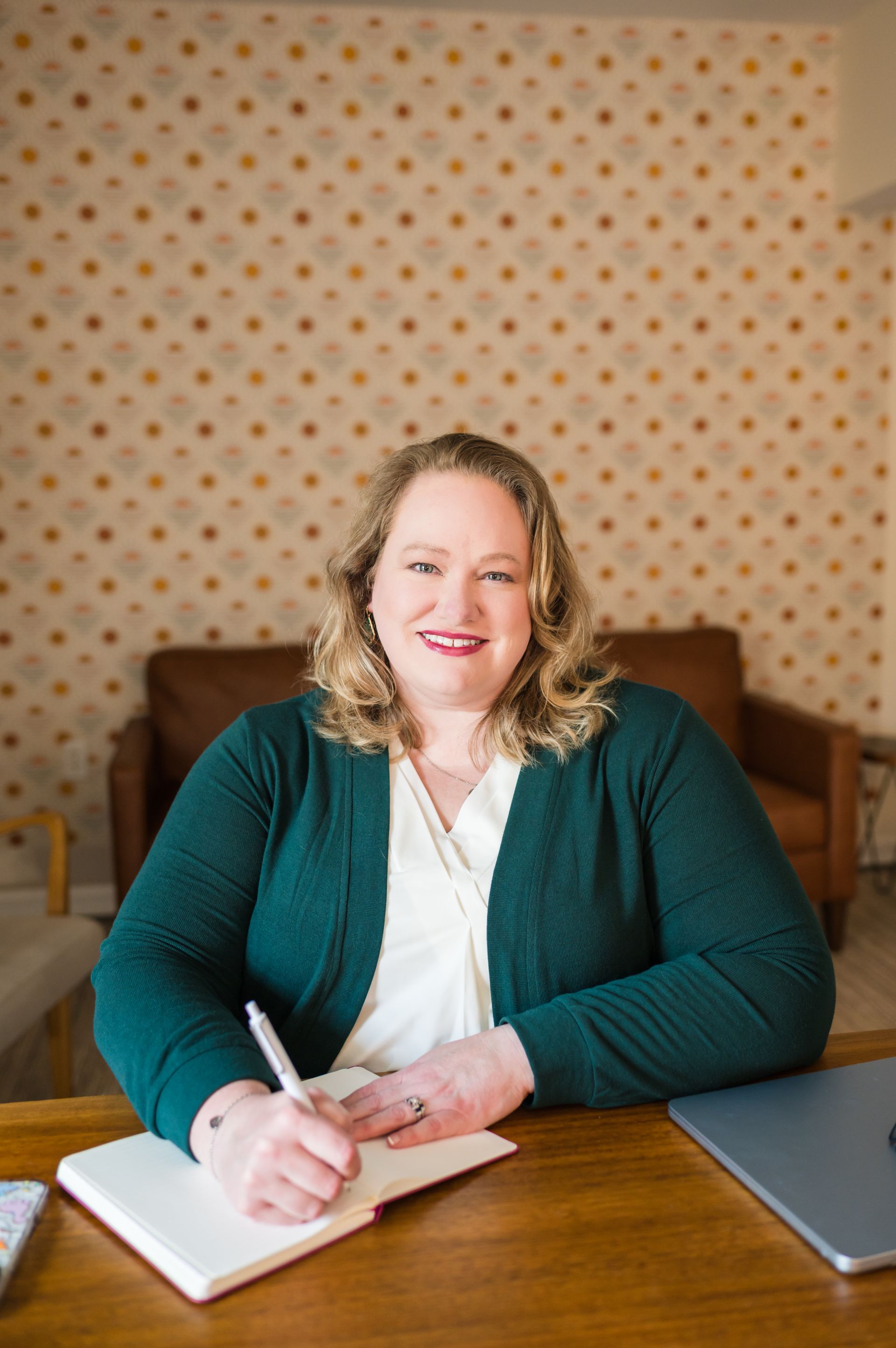 Woman with blonde hair, writing in notebook, smiling. Wearing green cardigan, sitting at desk.