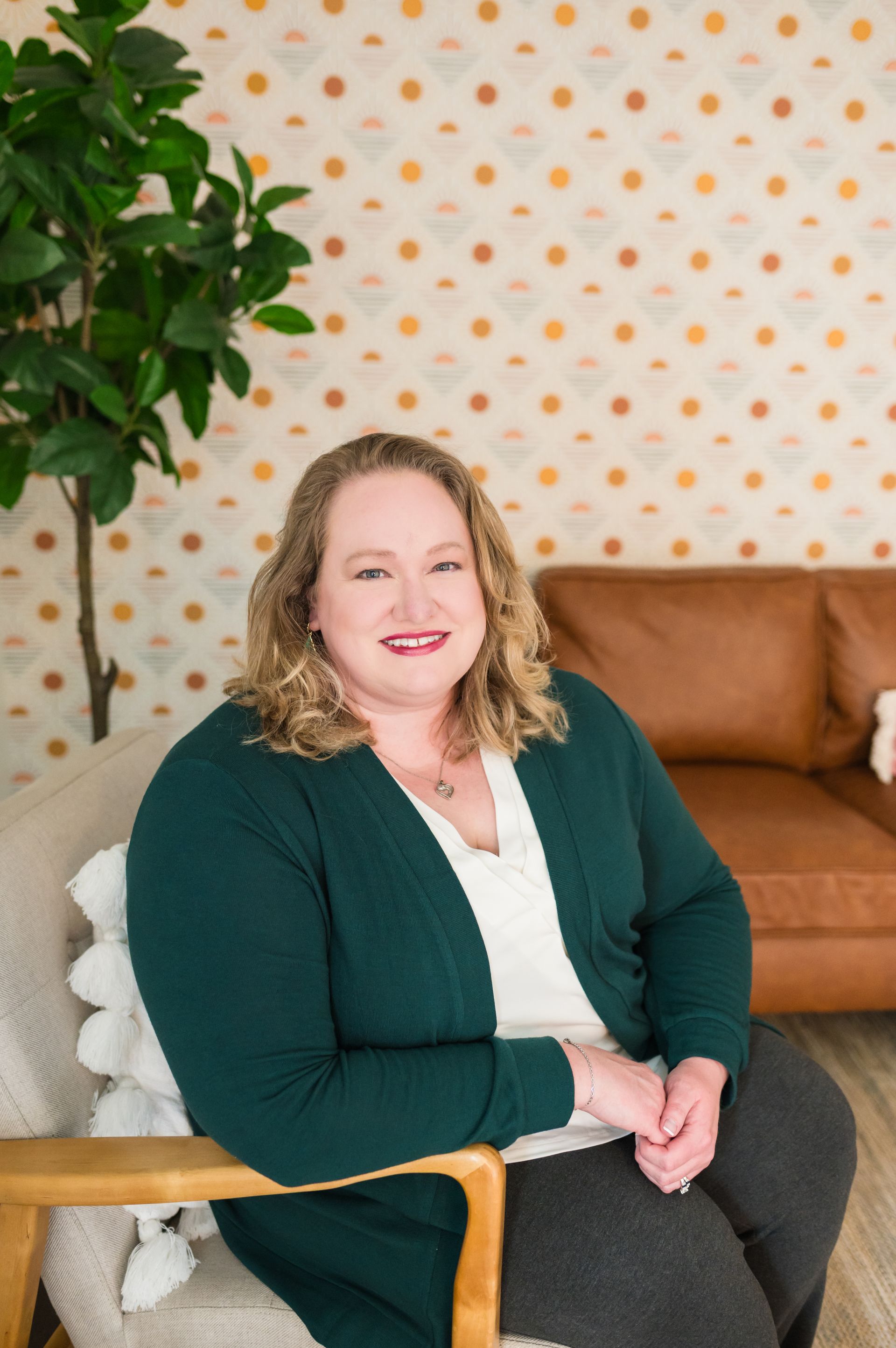 Woman with curly hair, wearing green cardigan, sits in a chair, smiling. Leather couch and patterned wall in background.