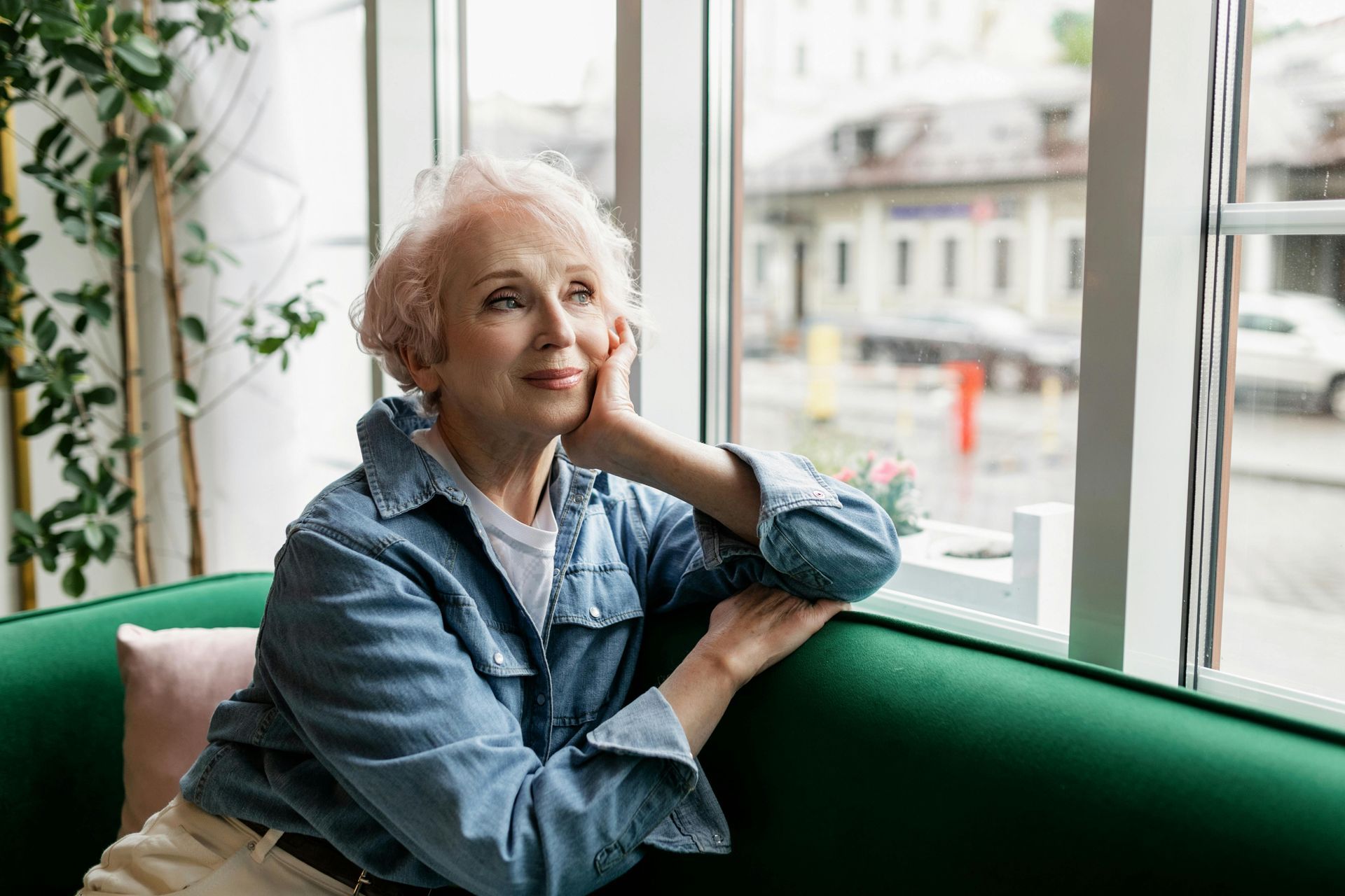 Woman in a denim shirt sitting on a green sofa by a window, looking outside with a thoughtful expression