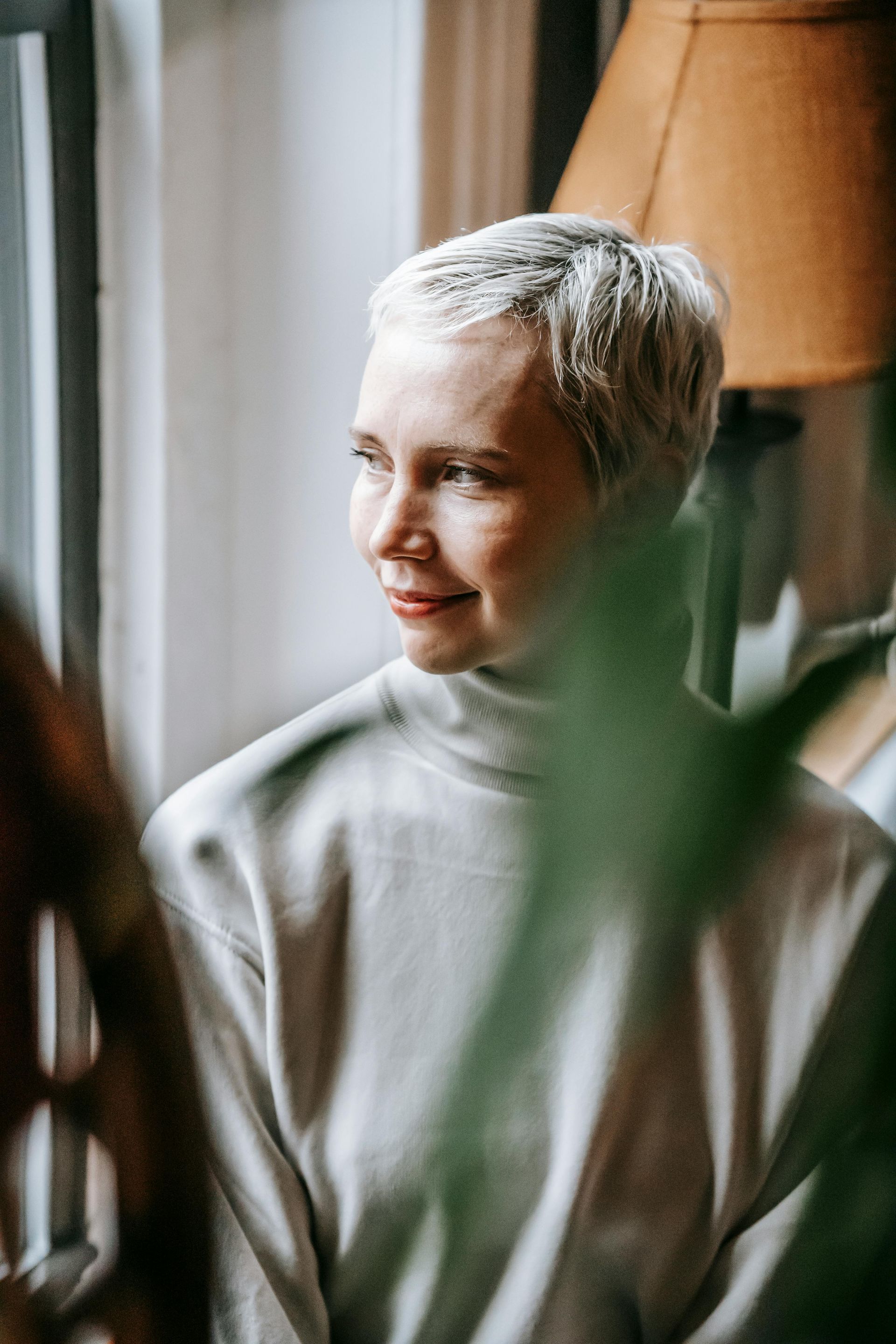 Portrait of a smiling woman in a light sweater by a window, with soft plants in the foreground