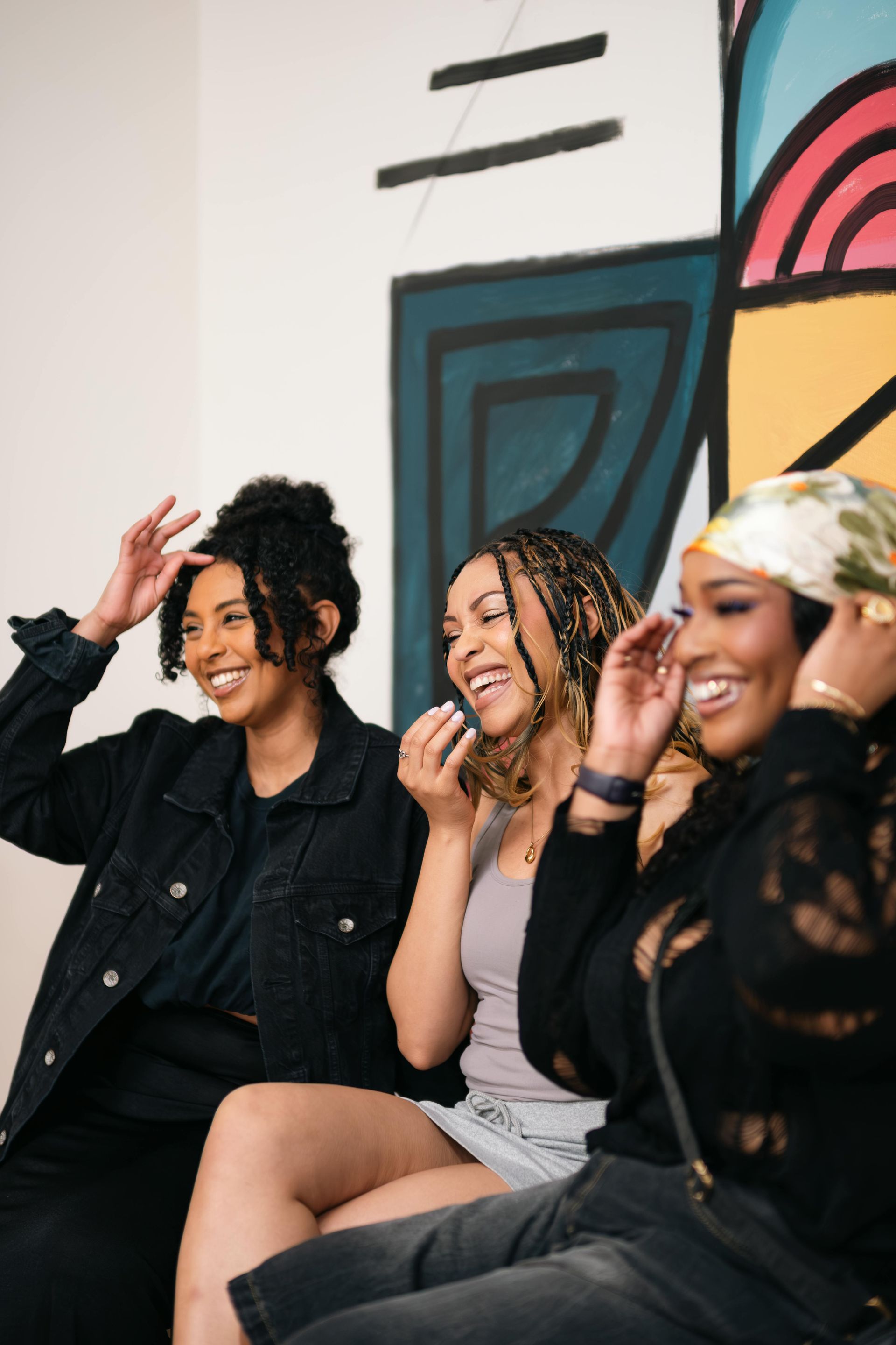 Three smiling friends sitting on a couch, laughing in front of a colorful mural.