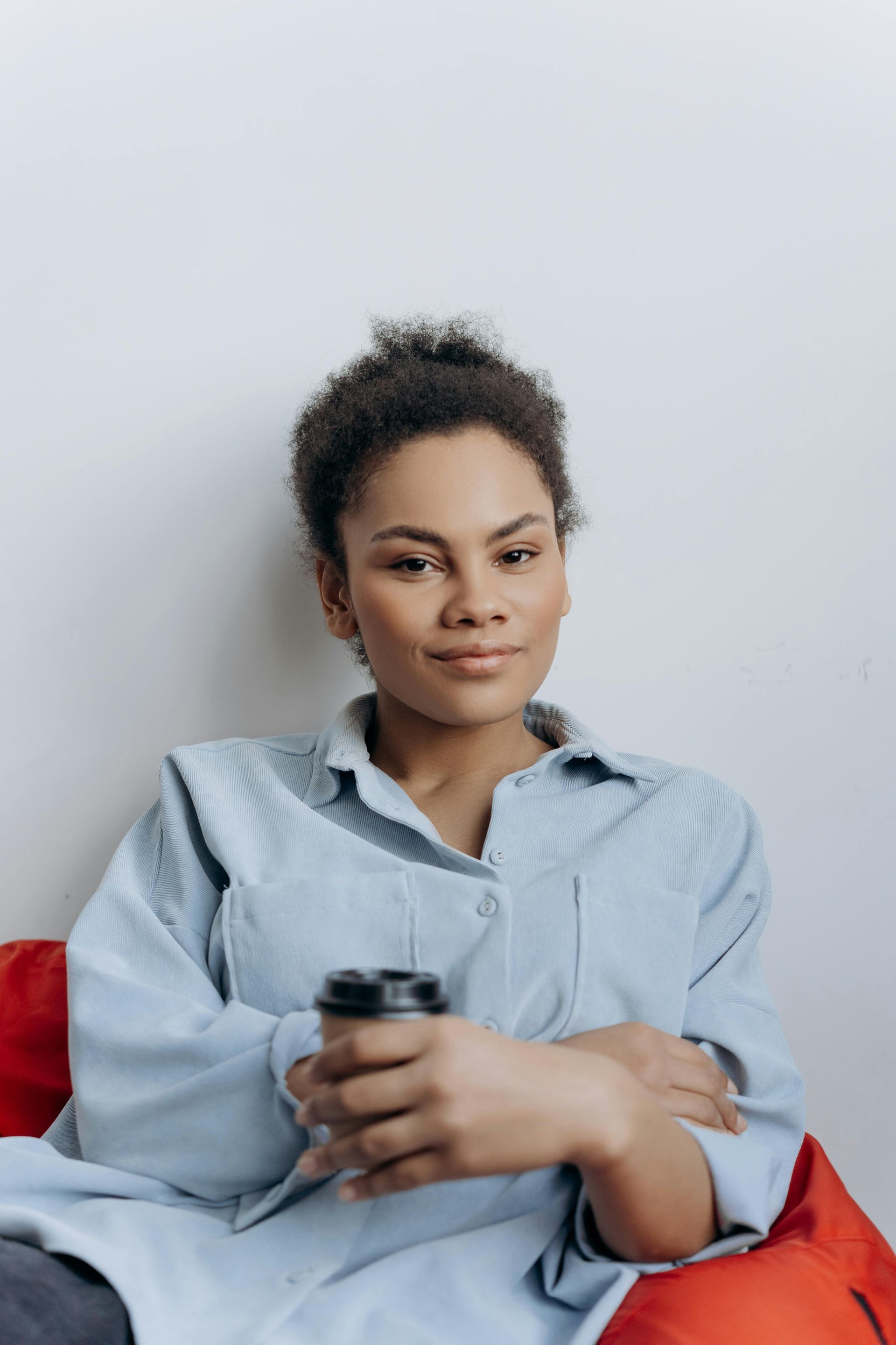 Woman in a light blue shirt seated on a red couch, holding a remote and looking at the camera.