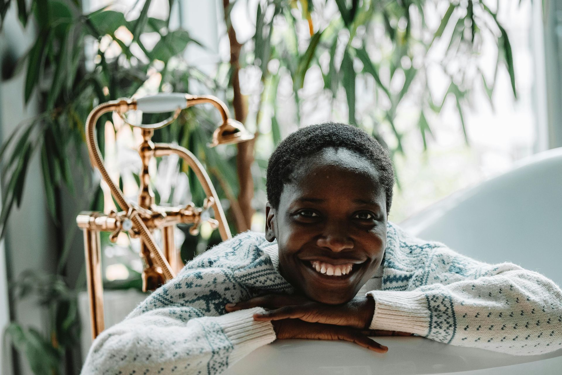 Smiling woman resting on a white bathtub edge in a bright bathroom with green plants and brass fixtures