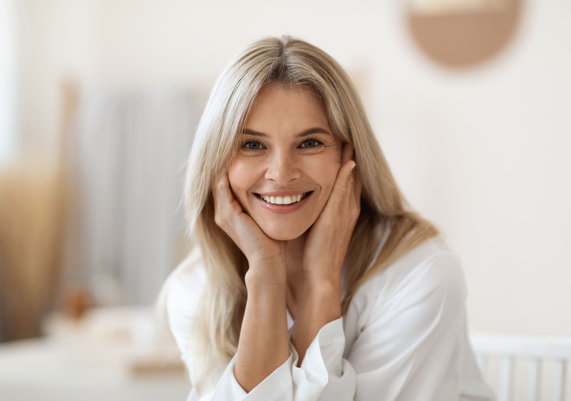 Blonde woman with hands on cheeks smiles brightly in a light-filled room.